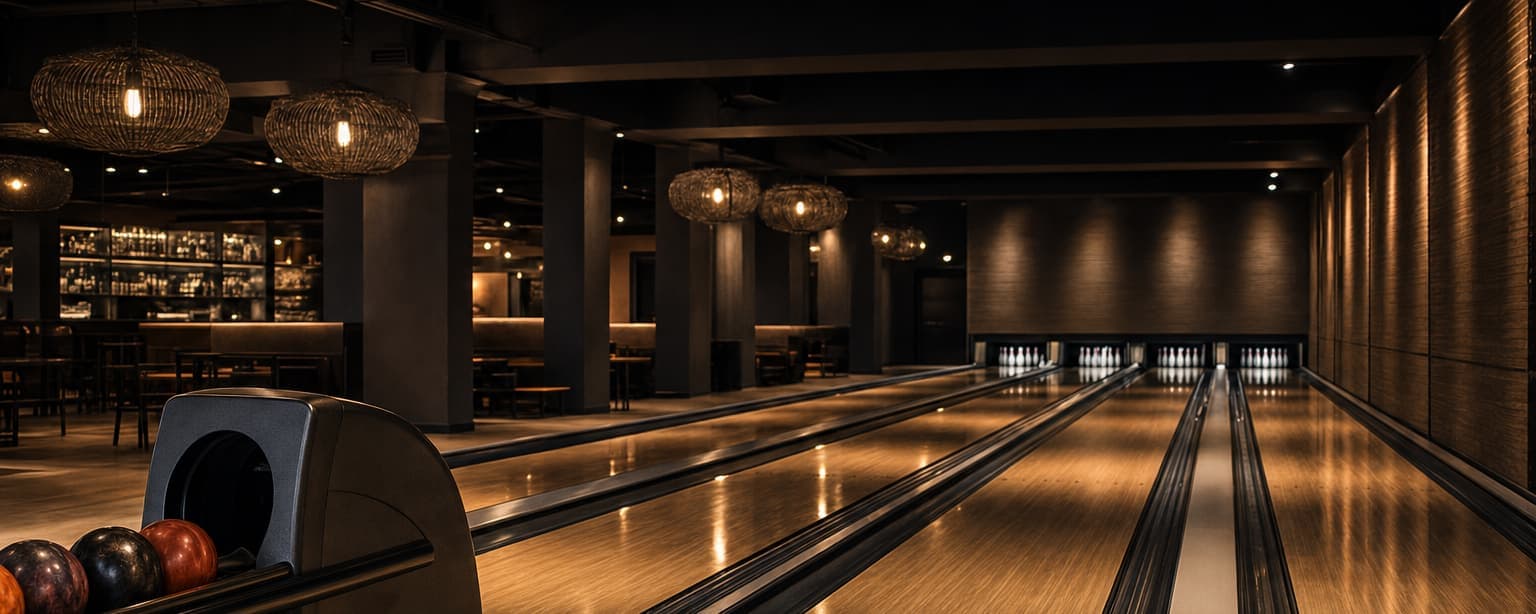A bowling center before opening, lanes lit down the row with ball returns and pin decks set, the bar and seating in shadow behind